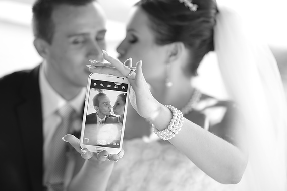 The bride is holding a mobile phone showing a photo of herself and the groom. They are sitting in a limo scene.
