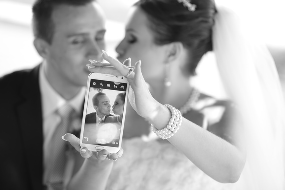 The bride is holding a mobile phone showing a photo of herself and the groom. They are sitting in a limo scene.