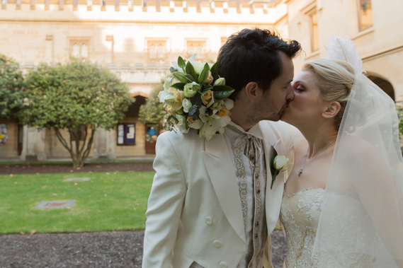 The bride is kissing her groom with the flowers around his neck. They are at Melbourne University, in Victoria. The groom is wearing a white and gold suit.