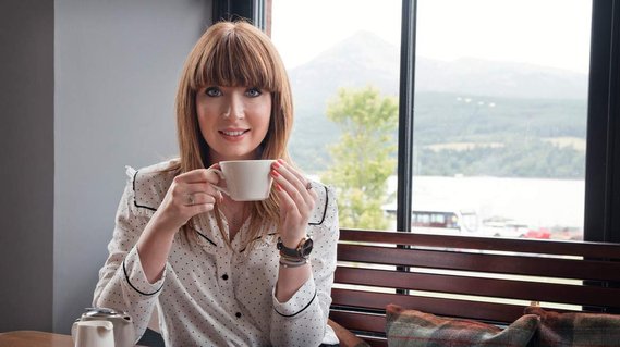 Nottingham-based branding photographer Karina Lyburn wears a polkadot shirt and sits with a cup of tea at a wooden table with Goat Fell through the window behind.
