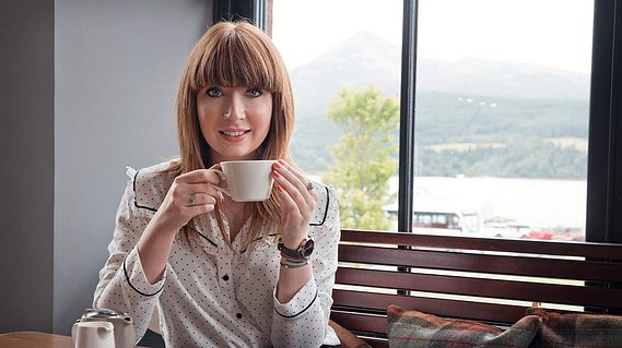 Nottingham-based branding photographer Karina Lyburn wears a polkadot shirt and sits with a cup of tea at a wooden table with Goat Fell through the window behind.