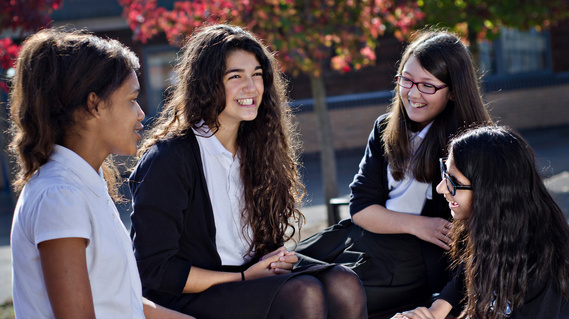 A group of schoolgirls wearing a black and white uniform sit laughing together on a bench in the sunshine.
