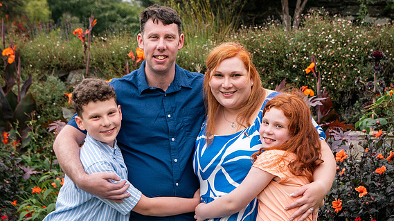 A family of four all wearing blue, white and orange hug in front of green bushes with orange flowers.