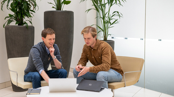 Two men sit at a table with a laptop between them and tall plants behind.