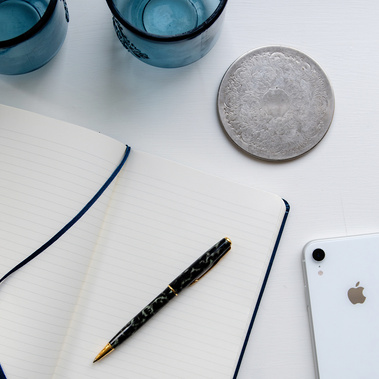 A flat lay photograph of an open journal, pen, white iPhone, silver coaster and blue glass jars.