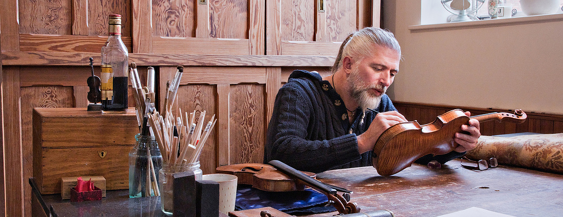 A man restores a violin sitting at a desk with paints and brushes on the desk.