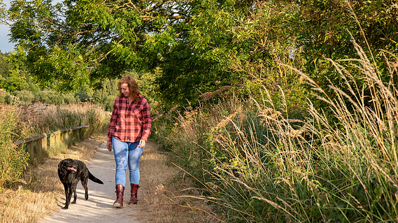 A woman wearing a check shirt and jeans walks along a path with a black labrador surrounded by long grasses and trees.