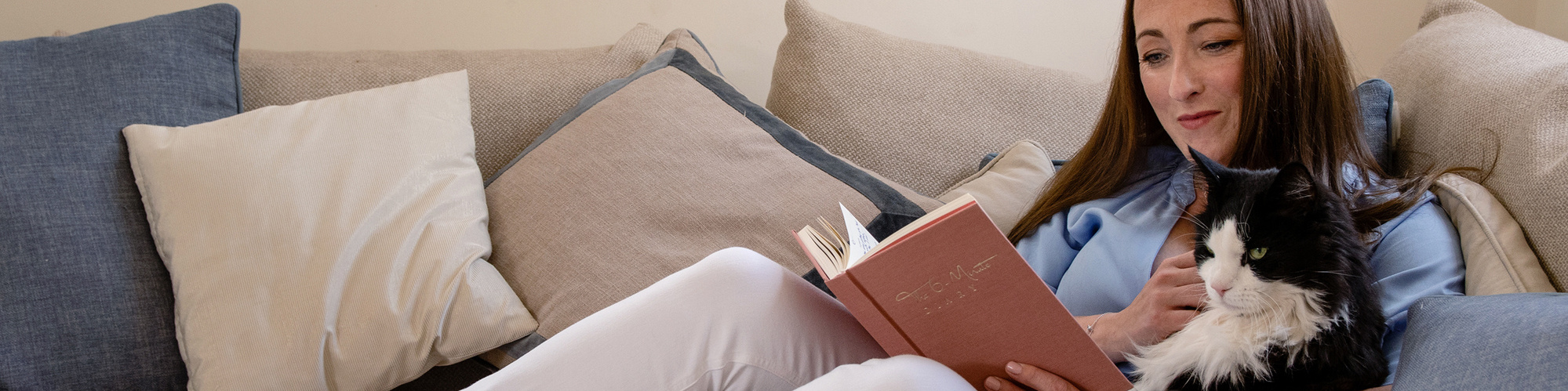 Interior design expert Natalie Gisborne wears a blue shirt, white jeans and sits on a cream corner sofa with her black and white cat reading a journal.