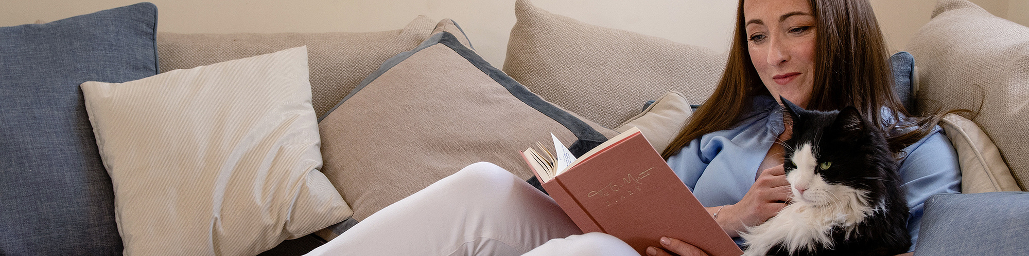 Interior design expert Natalie Gisborne wears a blue shirt, white jeans and sits on a cream corner sofa with her black and white cat reading a journal.