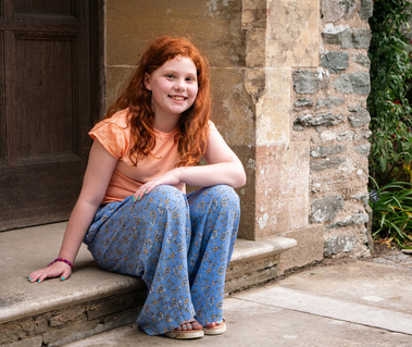 A girl with red hair wearing orange and blue sits on a step.