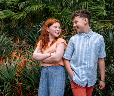A girl with red hair and arms crossed laughs with her brother, with green and orange plants behind.