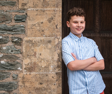 A boy wearing a blue shirt stands with arms crossed leaning against a stone pillar.