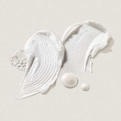 A close-up of two Deinde skincare formulations swiped across a neutral surface, highlighting the smooth, rich textures and active ingredients. Small droplets complement the composition, emphasizing hydration and product absorption. 