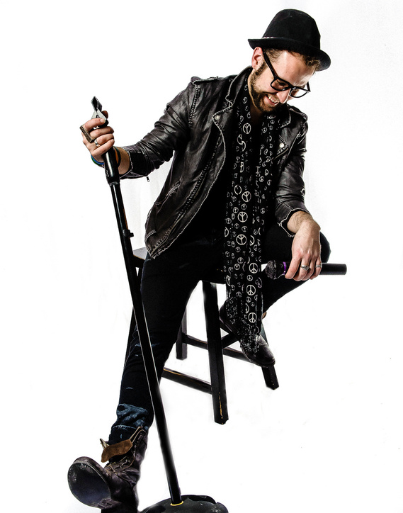 A man in black attire on a stool, leaning forward with his elbow resting on his knee. He holds a microphone stand with one hand. He is smiling and wears a hat, glasses, a leather jacket, and a patterned scarf.