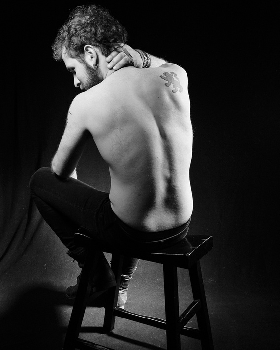A shirtless man sits on a stool with his back to the camera, his left hand resting on the back of his neck. He has a tattoo of a lion on his right shoulder blade. The image is in black and white and the background is dark.