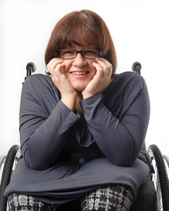 A woman with short brown hair and glasses is sitting in a wheelchair. She is wearing a long-sleeved grey top and patterned pants. She leans forward with her hands resting on her cheeks, smiling warmly. The background is white.