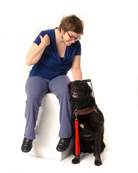 A person with short hair and glasses, wearing a blue top and light blue pants, sits on a white block, smiling and pointing to their black guide dog sitting next to them. The dog has a harness and a red leash. The background is plain white.