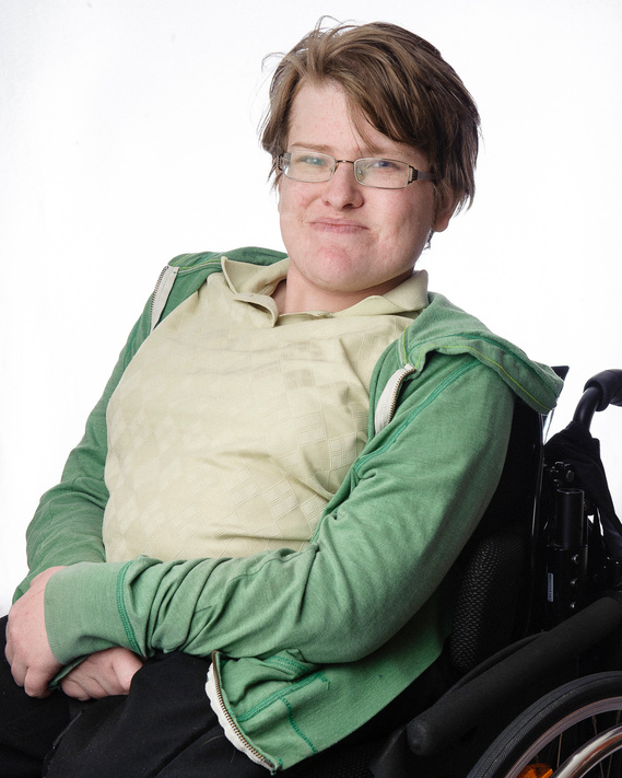 A young person with short light brown hair and glasses is sitting in a wheelchair. They are wearing a green hoodie over a light beige shirt and have their hands resting on their lap. The background is plain white.