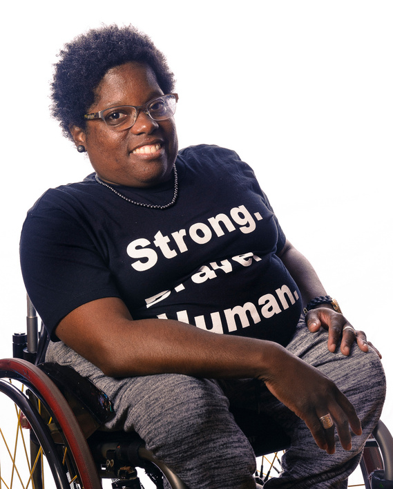 A person with dark skin and short curly hair, wearing glasses and a black t-shirt with the words 