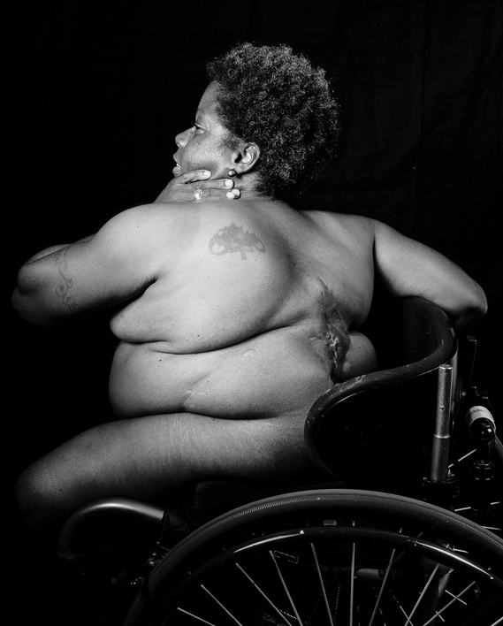 Black and white photo of a nude person of colour sitting in a wheelchair, with their back turned to the camera. The individual has visible scars on their back, short curly hair, and is touching their neck with one hand while looking to the side.
