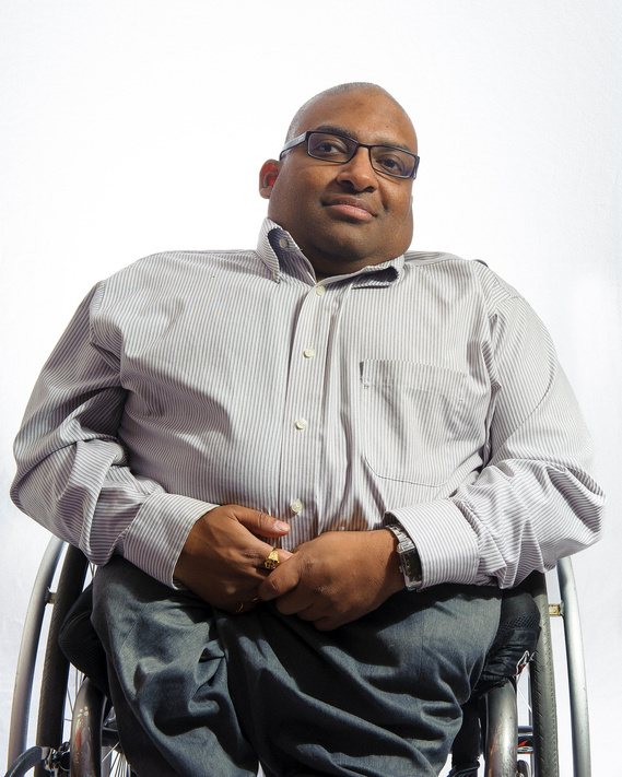 A man with dark skin wearing glasses and a calm expression is seated in a wheelchair. He is wearing a striped button-down shirt and dark pants. The background is plain and white.