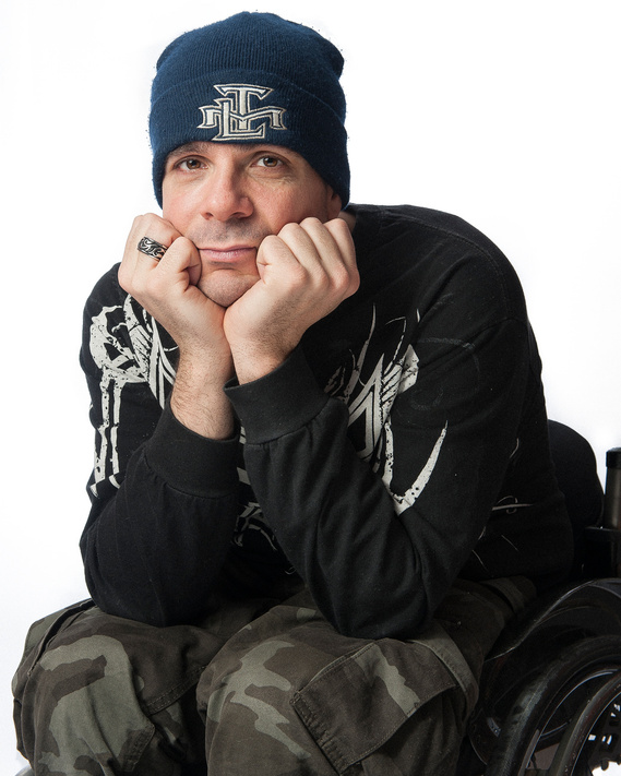 A person wearing a blue beanie and a black long-sleeve shirt with a white graphic design, rests their chin on their hands and looks forward. They are seated in a wheelchair and are also wearing camouflage pants snd a dark toque.