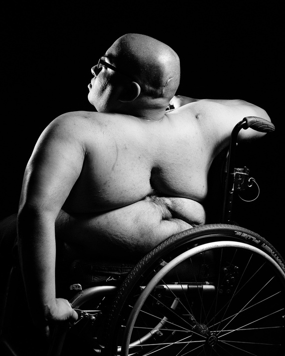 A black and white photo of a shirtless person sitting in a wheelchair. They are bald and wearing glasses, turned slightly to the side with their back to the camera. The lighting highlights the curves and contours of their body against a dark background.