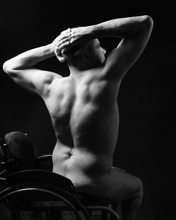 A black and white photograph of a nude person sitting in a wheelchair, viewed from the back. The person has their hands clasped behind their head, highlighting their muscular back and the contours of their body in dramatic lighting.