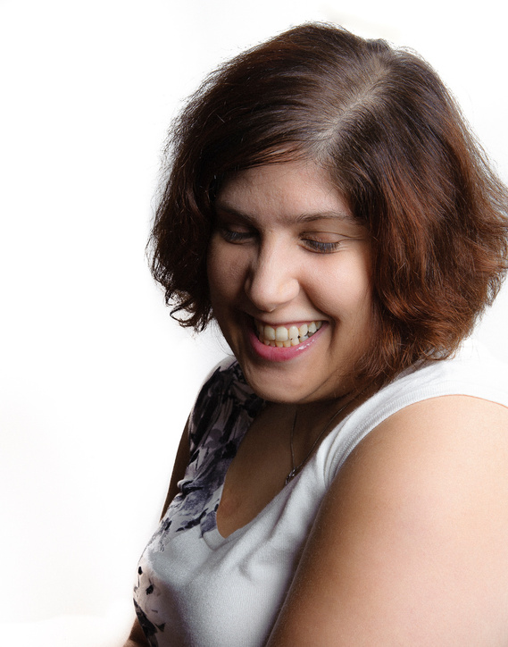 A person with shoulder-length brown hair is captured smiling downwards with their eyes closed. They have a joyful expression and are dressed in a white top with a floral pattern. The background is plain white, putting focus on their happy demeanor.

