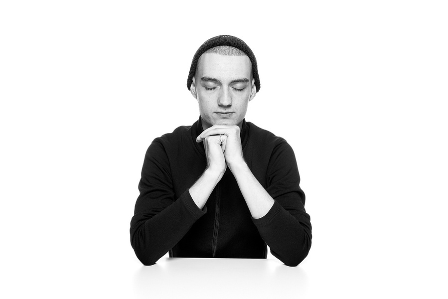  Ausdrucksstarkes Schwarz-Weiß-Portrait mit ruhiger, introspektiver Wirkung. Der junge Mann trägt eine Mütze und hält seine Hände gefaltet vor sich, während er die Augen geschlossen hat. Die minimalistische Ästhetik betont Emotion und Tiefe. Perfekt für k