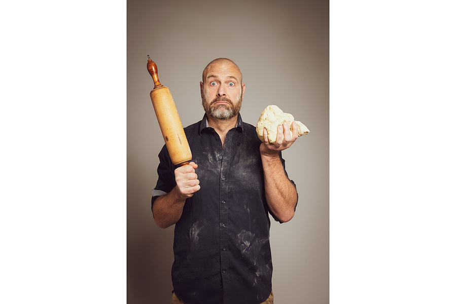 Dynamisches Portrait eines Bäckers mit Mehl auf Hemd, Teig in der Hand und Nudelholz. Der humorvolle Ausdruck vermittelt Handwerkskunst und Leidenschaft. Ideal für Werbekampagnen, Gastronomie-Branding oder Social Media Content.