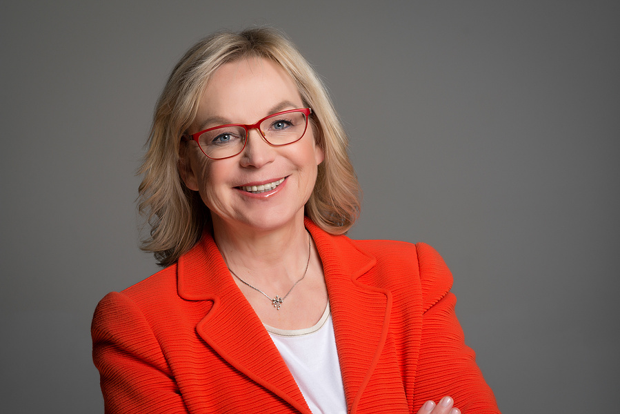 Freundliches Businessportrait mit starkem Ausdruck. Die Frau trägt einen roten Blazer und eine markante Brille, während sie selbstbewusst vor neutralem Hintergrund posiert. Perfekt für LinkedIn, Unternehmenswebsites oder PR-Auftritte.