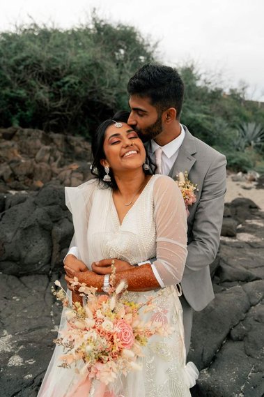 Des mariés se prenant dans les bras sur une plage de La Réunion avec un bouquet rose. 