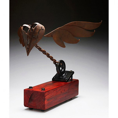 A large steel and wood sculpture of a winged heart attempting to fly but held by a chain to the wooden base