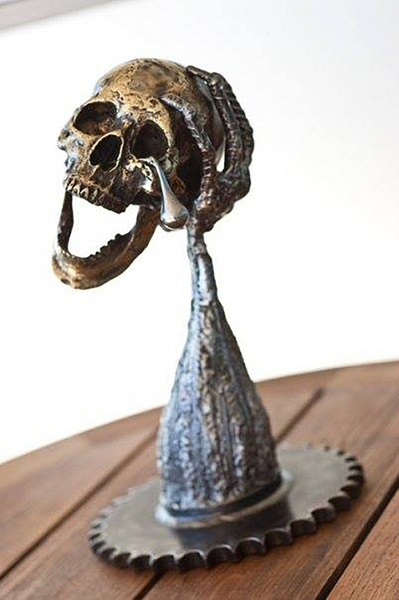 A steel and brass sculpture of an arm with a brass skull resting on the hand with a single steel tear dripping from an eye socket