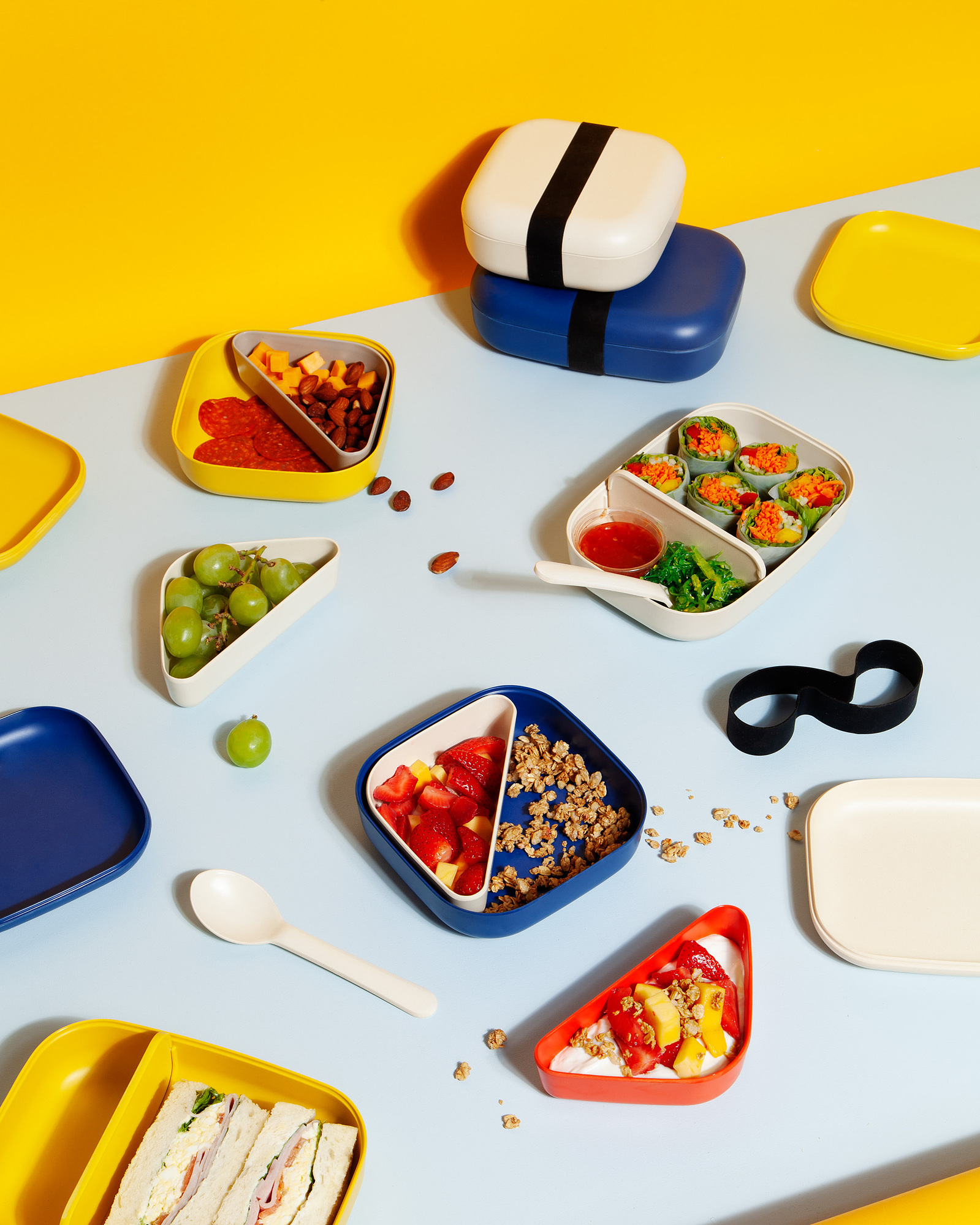 Bright commercial food styling on a blue tabletop with a yellow background, multiple bento boxes, dishes and flatware are scattered and styled on the tabletop with various food props