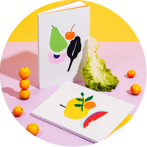 Photography and Styling of stationery notebooks by photographer stylist Becca M. Image of two notebooks with fruit and vegetable illustration designs, one standing on laying, with kumquats and a piece of lettuce as props on a lavender and yellow backdrop.