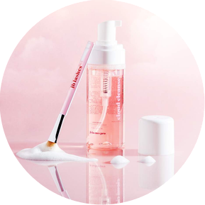 Product Photography and Styling for Beauty cleanser by photographer stylist Becca M. Image of a pink foam cleanser bottle on a pink background, with cleaning brush leaning against the product with a puddle of foam. Pink with clouds in the background.