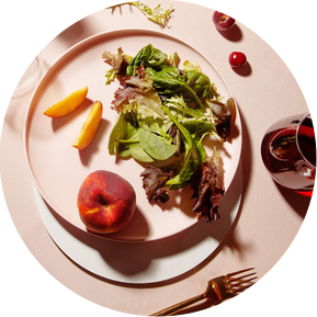 Photography and food tabletop styling by photographer and prop stylist Becca M, image of a pink table backdrop with pink pastel plates from target with a peach spring mix salad and scattered cherries and glasses of wine in the corner of the photo.