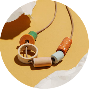 Product Photography and styling by photographer stylist Becca M of a beaded handmade clay necklace with warm colors and different shapes, laying out on a warm yellow background with torn paper around the edges framing the jewelry. 