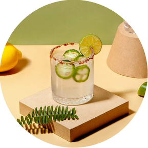 Photography and prop styling by photographer stylist Becca M. Product lifestyle of a spicy jalapeño margarita in a rocks glass on a brown coaster. tall empty glasses balanced in the background. Lemon, jalepeno and leaf as  props / garnish on green/yellow