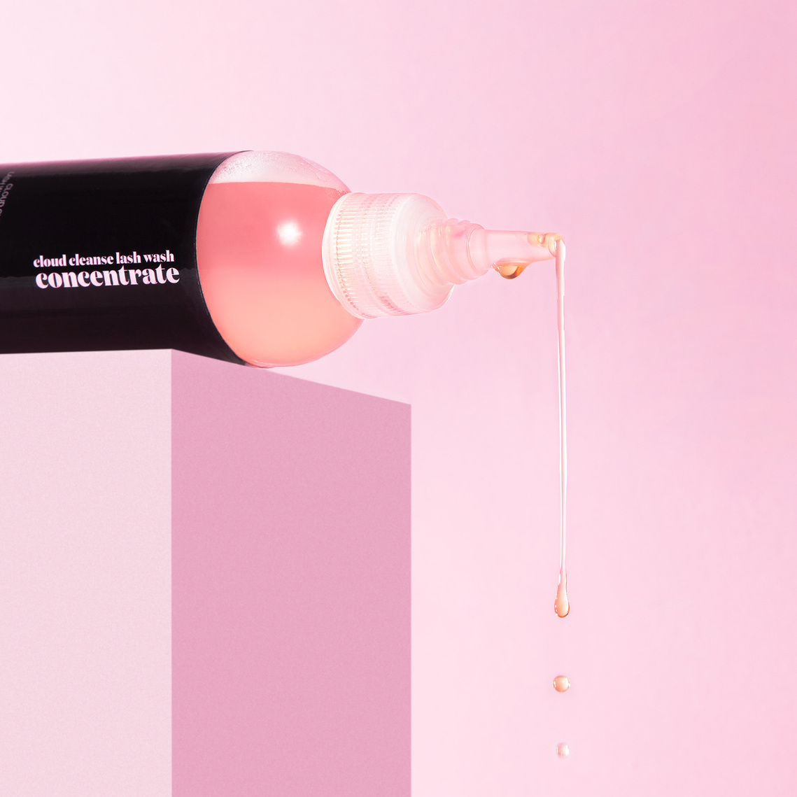 JB Lashes Pro Cloud Cleanse Concentrate Wash on pink background with drip coming out of tip of bottle on a pink platform close up product photography prop styling by photographer stylist Becca M los angeles