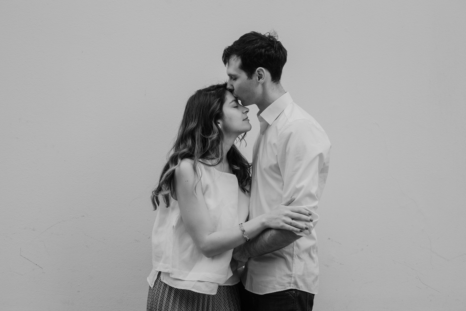 man kisses his girlfriend's forehead in front of a white wall