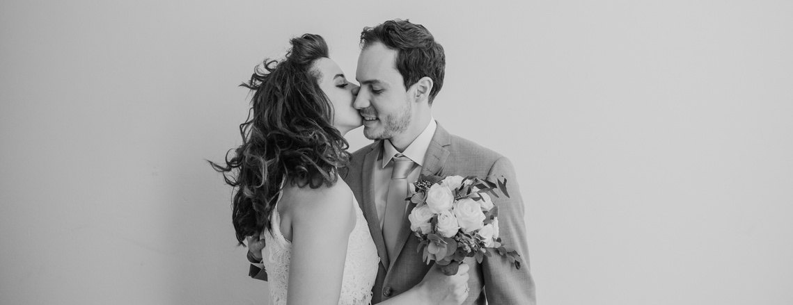 Newly married couple kissing each other on the cheek. The woman holds a bouquet of flowers in her hand. 