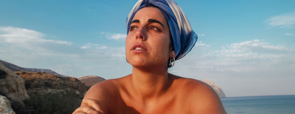 Woman with blue headscarf cries as she sits on the beach watching the sunset