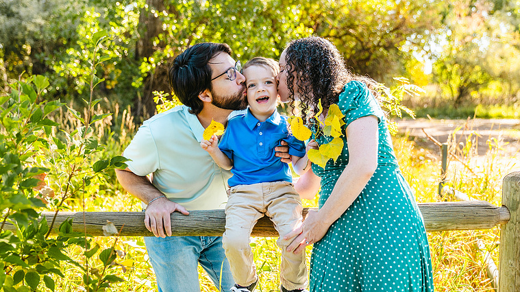 A young boy dressed in blue sits on a split rail fence while his mom and dad both kiss him on the cheek, the boy is smiling and holding leaves during a family photo session in Fort Collins, Colorado.