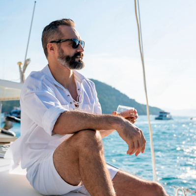 Image of middle-aged man wearing sunglasses holding a drink whilst sitting on yacht in the sunshine