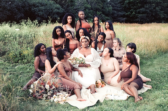 a group of bridesmaids sit surrounding a bride in a field with flowers