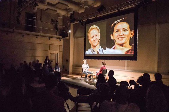 a man and woman sit across from each other on stage with close up video feeds of their faces projected to an audience.