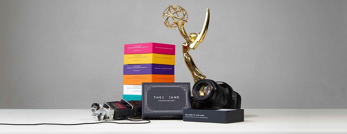 The And card game is presented on a white table in front of a stack of various colored editions, an Emmy award trophy, a DSLR camera, and an audio recorder.
